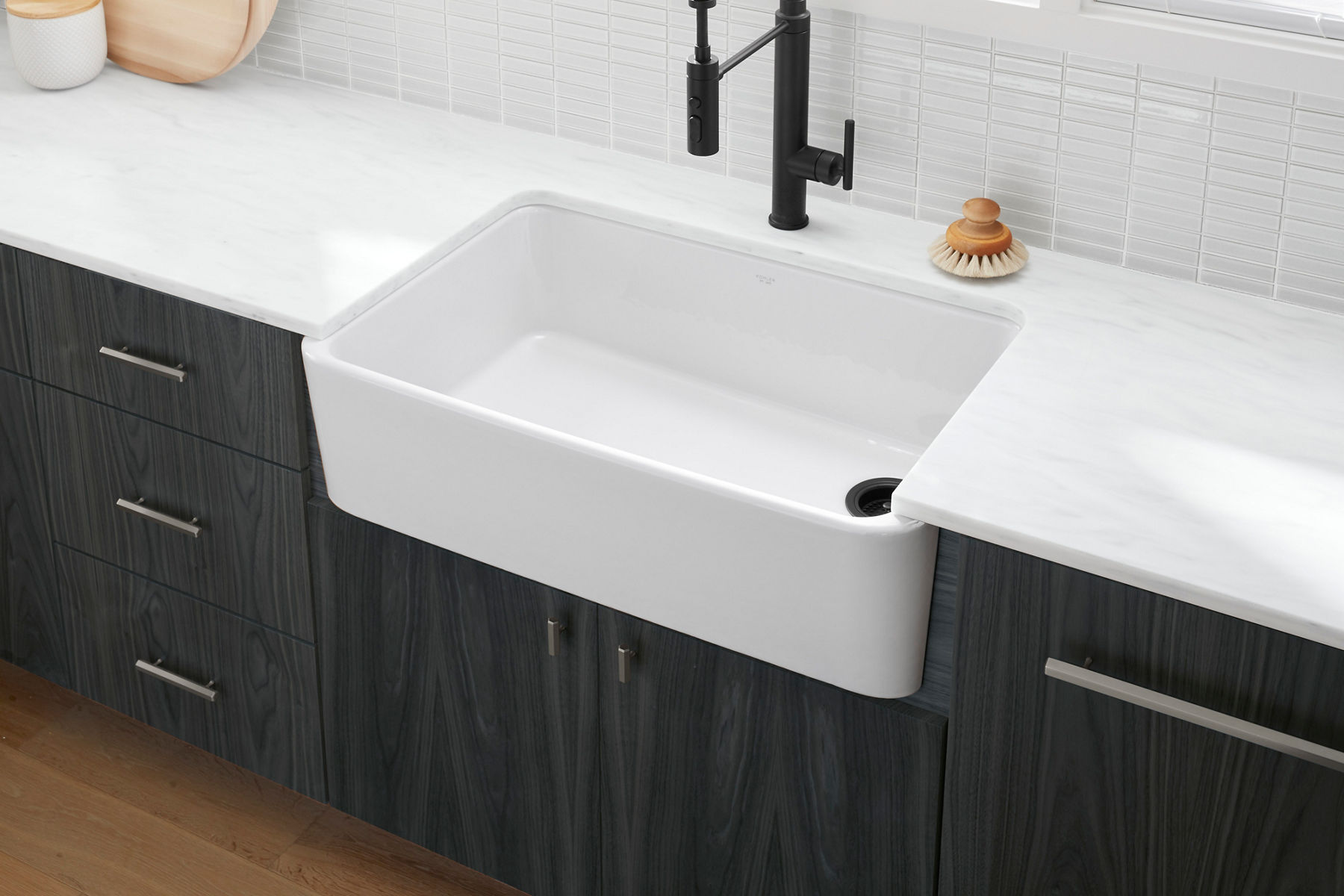 A close-up of a white farmhouse sink with a black faucet set in a modern kitchen with dark wood cabinets and a white tiled backsplash.