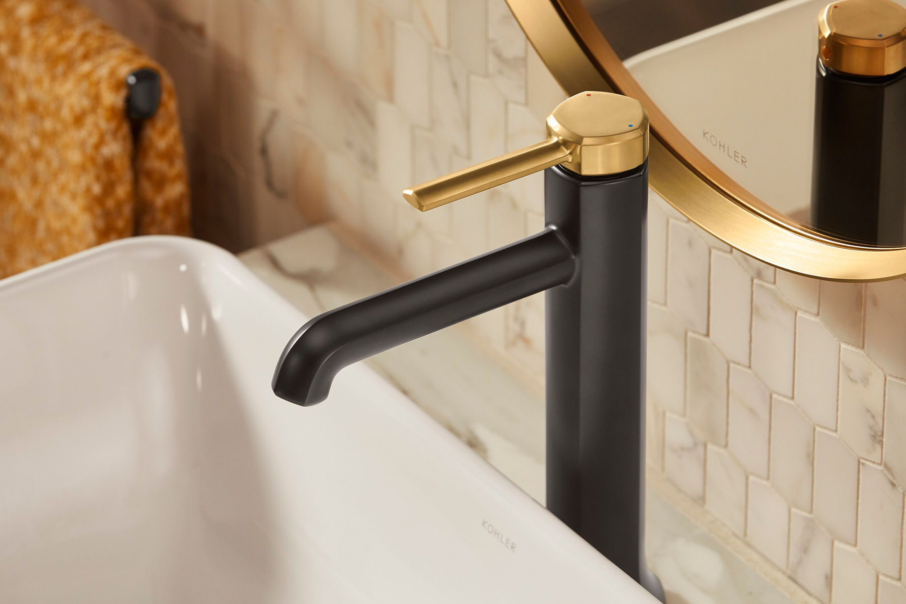 A close-up of a modern bathroom faucet with a black spout and gold handle, installed on a white sink with a marble backsplash.