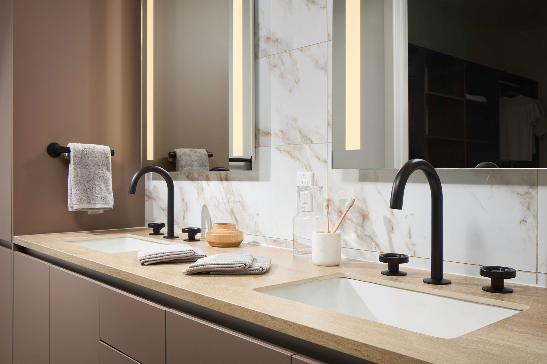 A contemporary bathroom with a double vanity in a brown color with a light brown countertop, white undermount sinks, black widespread faucets, and a white stone wall with brown marbling with two internally lit mirrors.