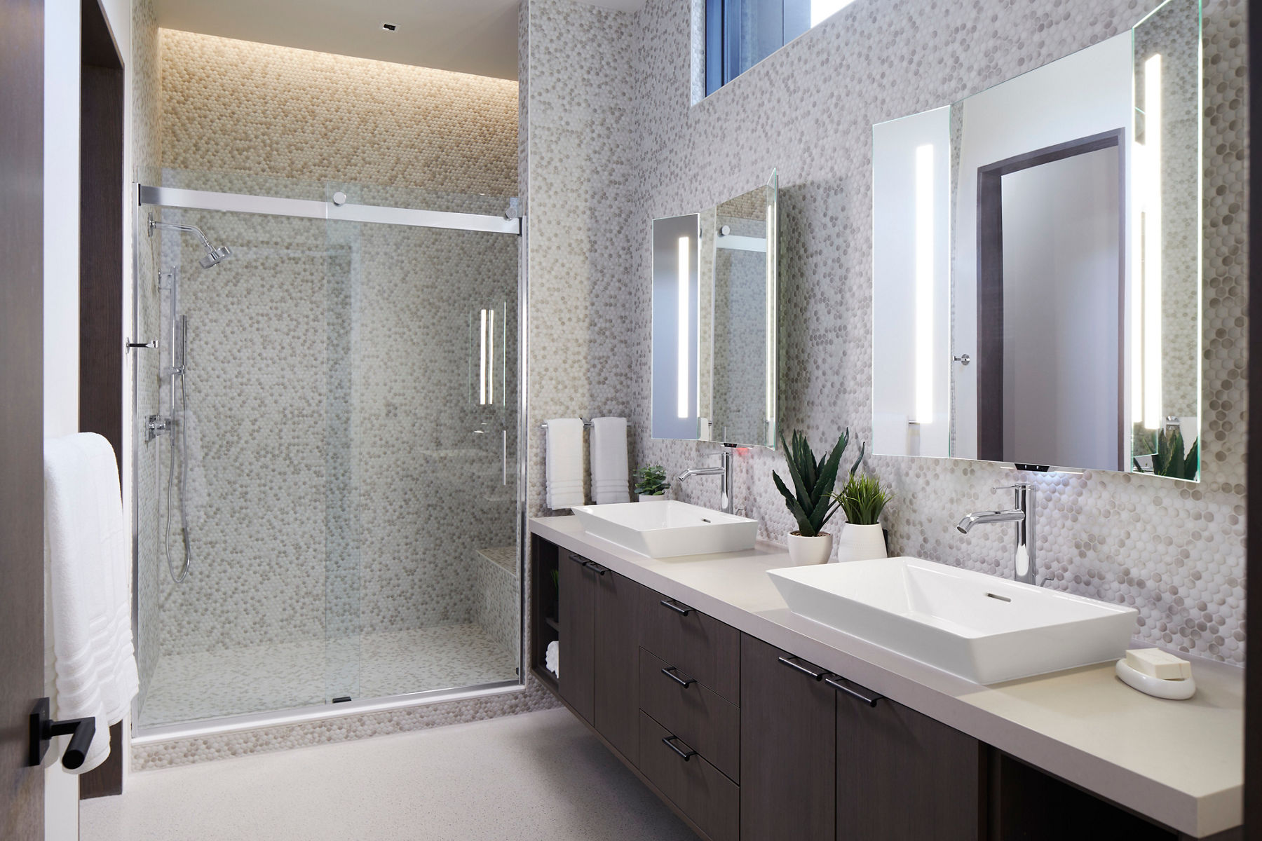 A bathroom with a double vanity in dark wood, white vessel sinks, chrome single handle faucets, internally lit mirrors, and a shower area with a sliding glass door.