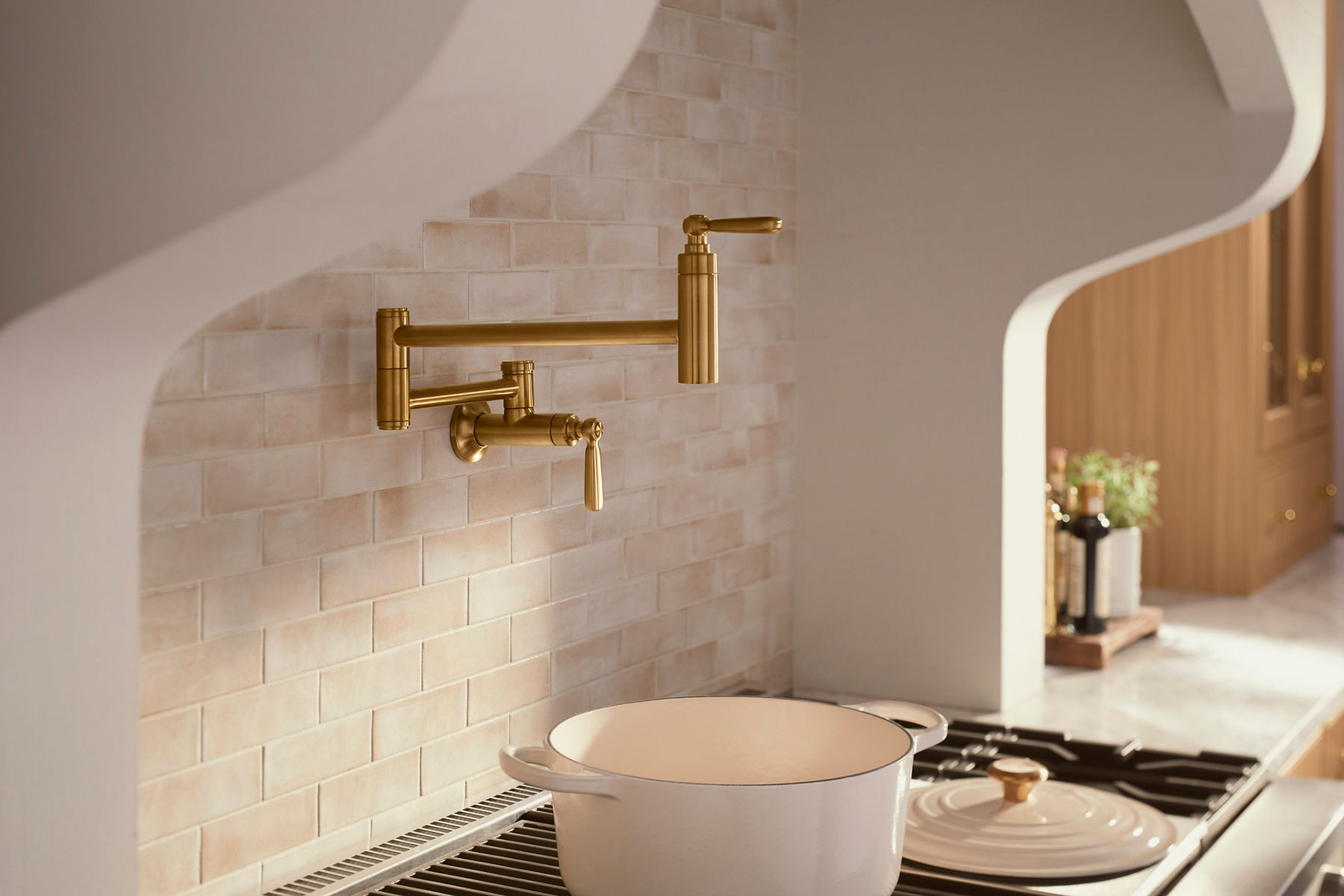 A luxurious kitchen featuring a gold wall-mounted pot filler faucet above a stove with a large cream-colored Dutch oven. The backsplash consists of light beige subway tiles, and the space is designed with a soft, warm color palette. The arched hood adds an elegant touch, framing the cooking area beautifully.