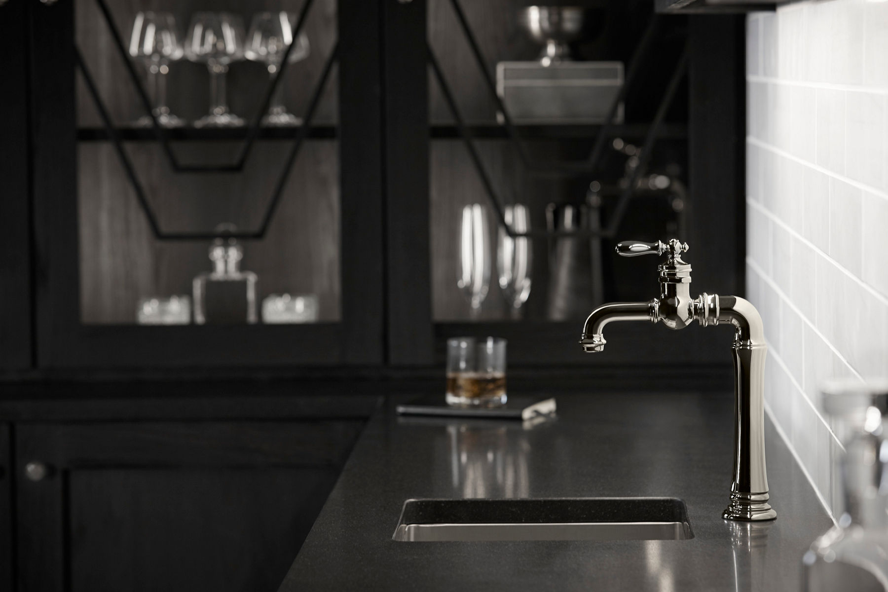 A sophisticated kitchen or bar area featuring a vintage-inspired polished chrome faucet with intricate detailing. The faucet is mounted on a dark countertop with an undermount sink. In the background, there is a glass of whiskey and a dark cabinet with glassware, creating a moody and elegant atmosphere. The white subway tile backsplash contrasts with the dark tones, adding a touch of brightness to the space.