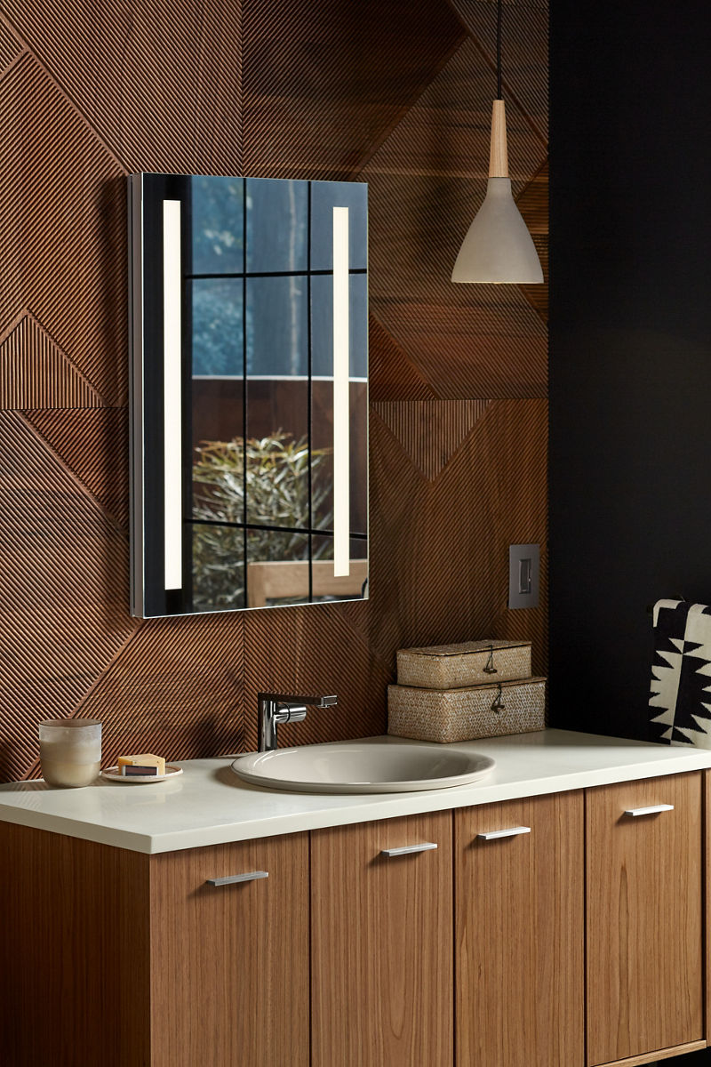 A recessed, lighted medicine cabinet against a wood wall engraved with large geometric shapes.