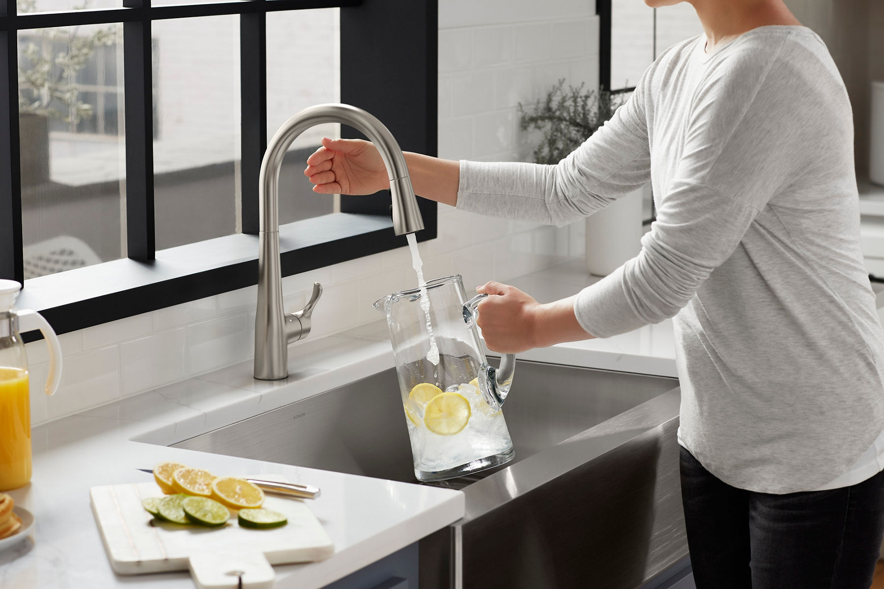 A person filling a glass pitcher with water and lemon slices from a modern kitchen faucet set against a white tiled backsplash and a large window.