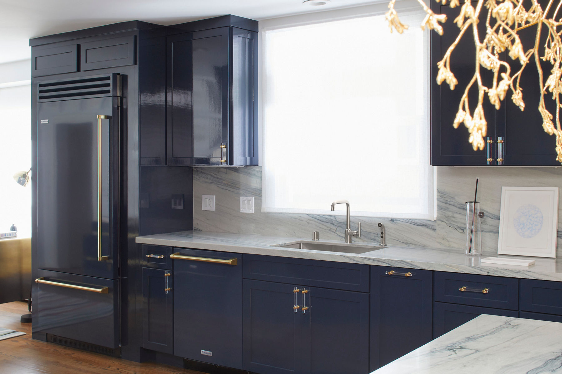 A modern kitchen with a sleek stainless steel faucet and an undermount sink set into a luxurious marble countertop with a matching marble backsplash. The cabinetry is painted a rich navy blue, accented with gold hardware. A large window above the sink allows natural light to brighten the space, emphasizing the clean and elegant design.