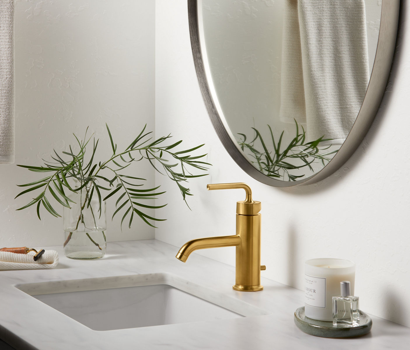 Un dessus de meuble-lavabo blanc de salle de bain avec plateau pour accessoires, un vase contenant des branches et un robinet à poignée à levier unique KOHLER Purist en laiton brossé moderne Vibrant.