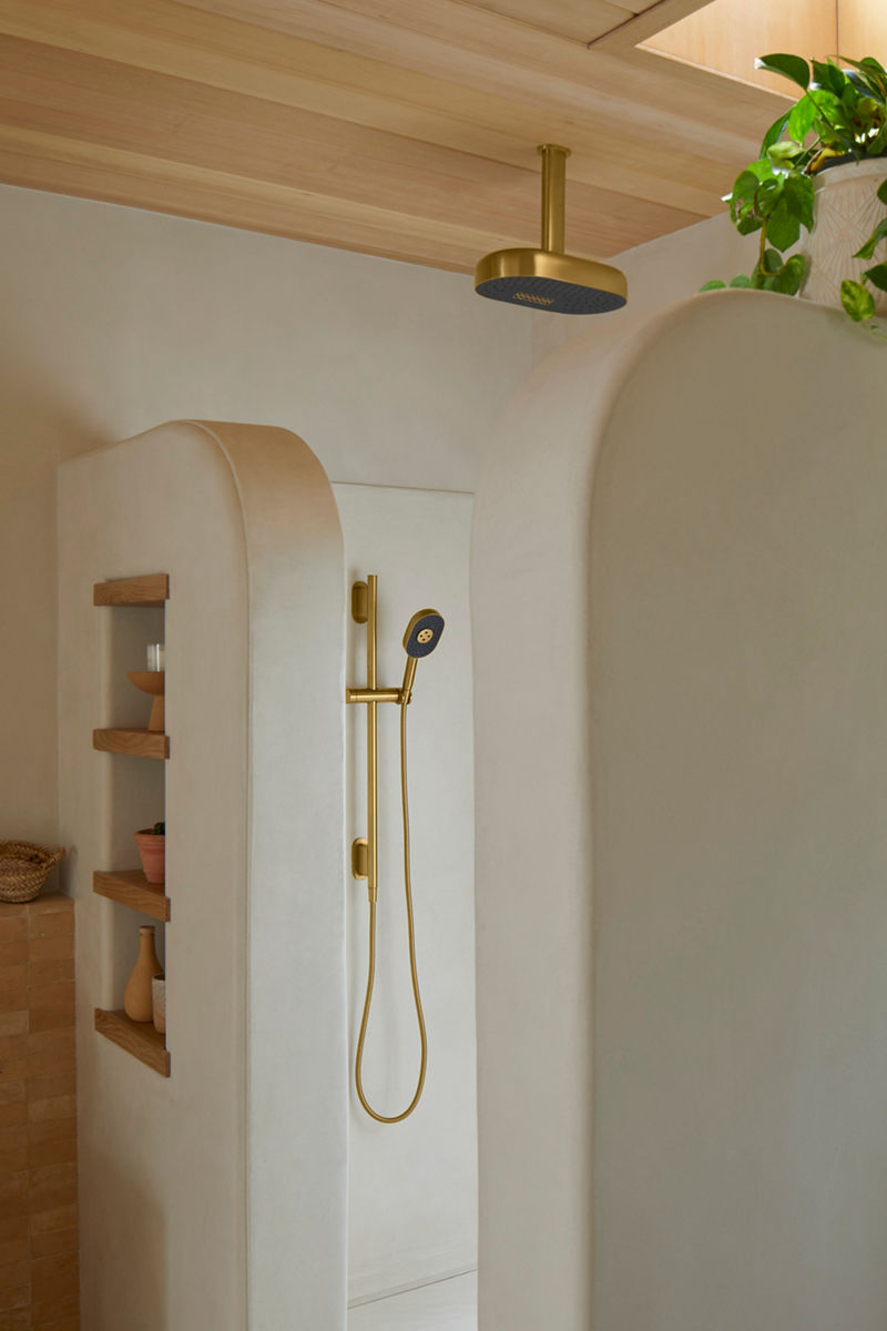 Image of the shower area in the primary bathroom, featuring a Kohler Statement rainhead showerhead coming down from the ceiling, plus a handshower spray and adjustable bar in matching brass finish. The shower walls are a light terracotta plaster finish, replicating the colors and texture found in the southwest desert around their Tuscon, Arizona, home. 