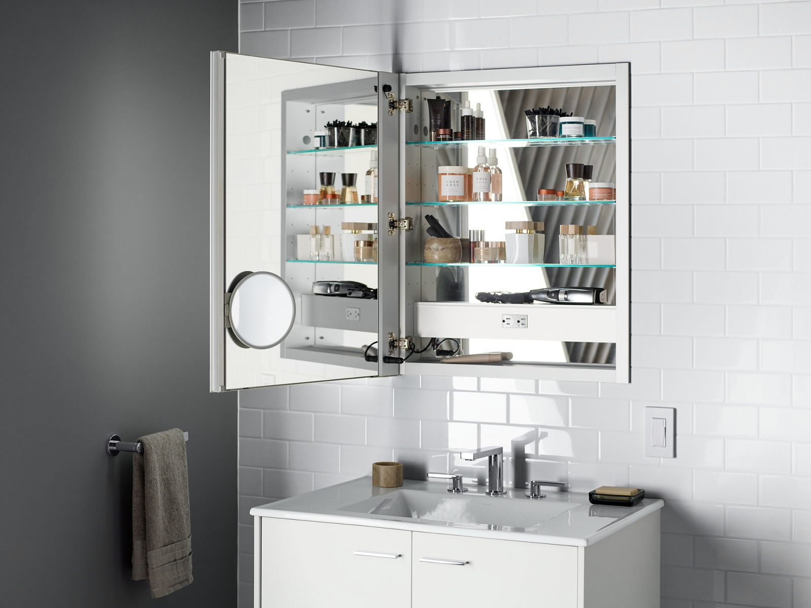 A medicine cabinet with its door open to show a mirrored inside door and back panel, along with a magnification mirror, an electrical outlet, and storage for numerous daily items, all set against a white tile wall and over a white vanity.