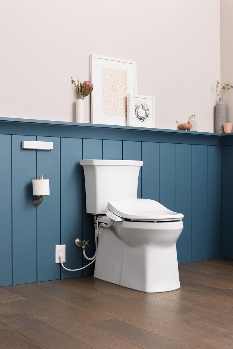 A bathroom with blue wainscoting, a shelf with a vase of flowers and framed artworks, and a white KOHLER elongated toilet with bidet seat.