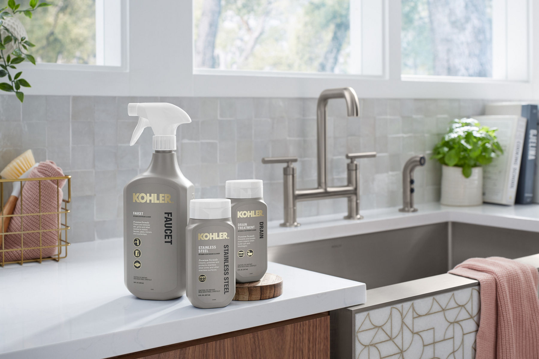 A spray bottle and two jars of KOHLER cleaning products sit on a white counter, next to a sink with a deck-mounted bridge faucet.