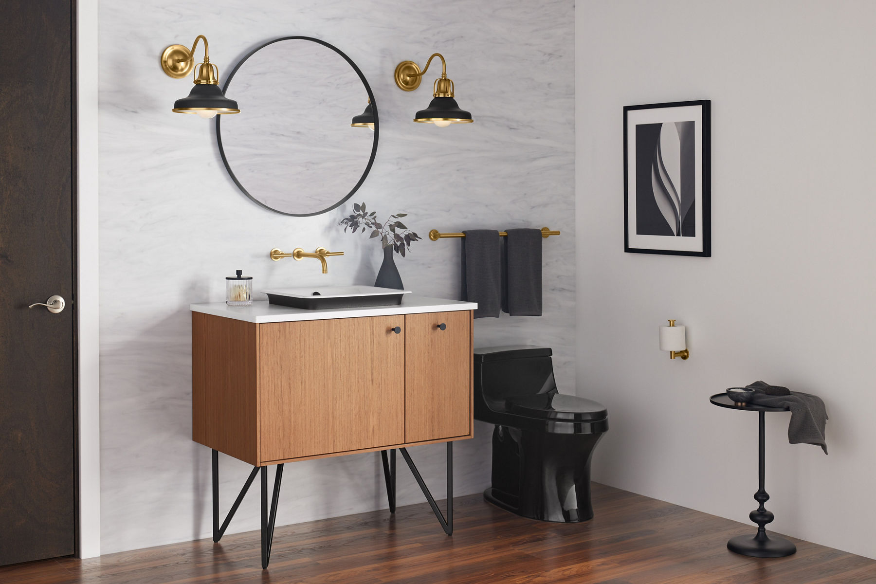 A round, black-framed mirror on a marbled gray wall above a wood-grain vanity with a white vessel sink and brushed brass faucet and accessories.