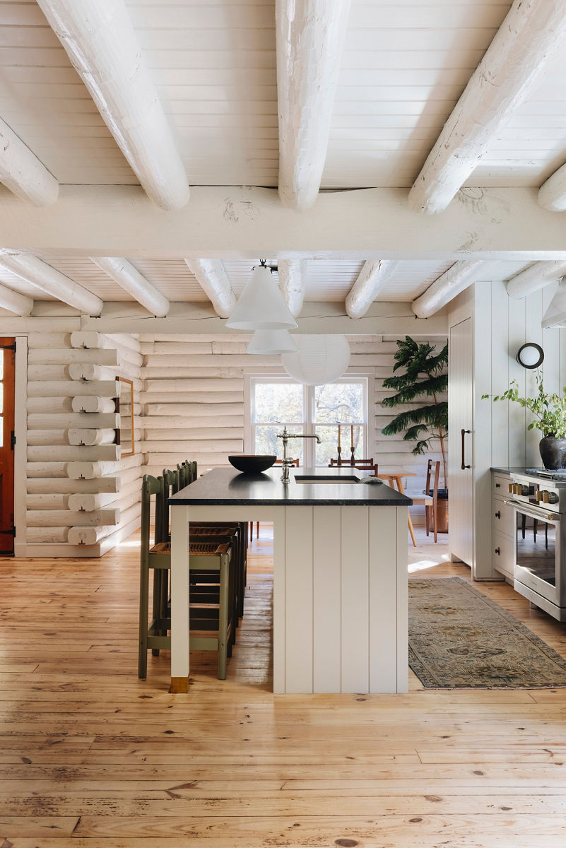 Wide image looking down the long way of the kitchen island, seeing the white wood base island seating area. The profile of the Kohler Artifacts faucet is centered on the island and between the sun-filled windows in the distance. Painted white logs of the cabin are visible on the walls and the ceiling. 