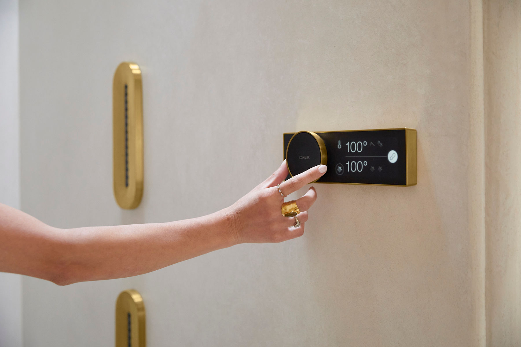 Close-up image of a hand adjusting the dial on the Kohler Anthem shower control panel. The panel face is black and the trim is brass, which matches the shower sprays that can be seen in the distance. 