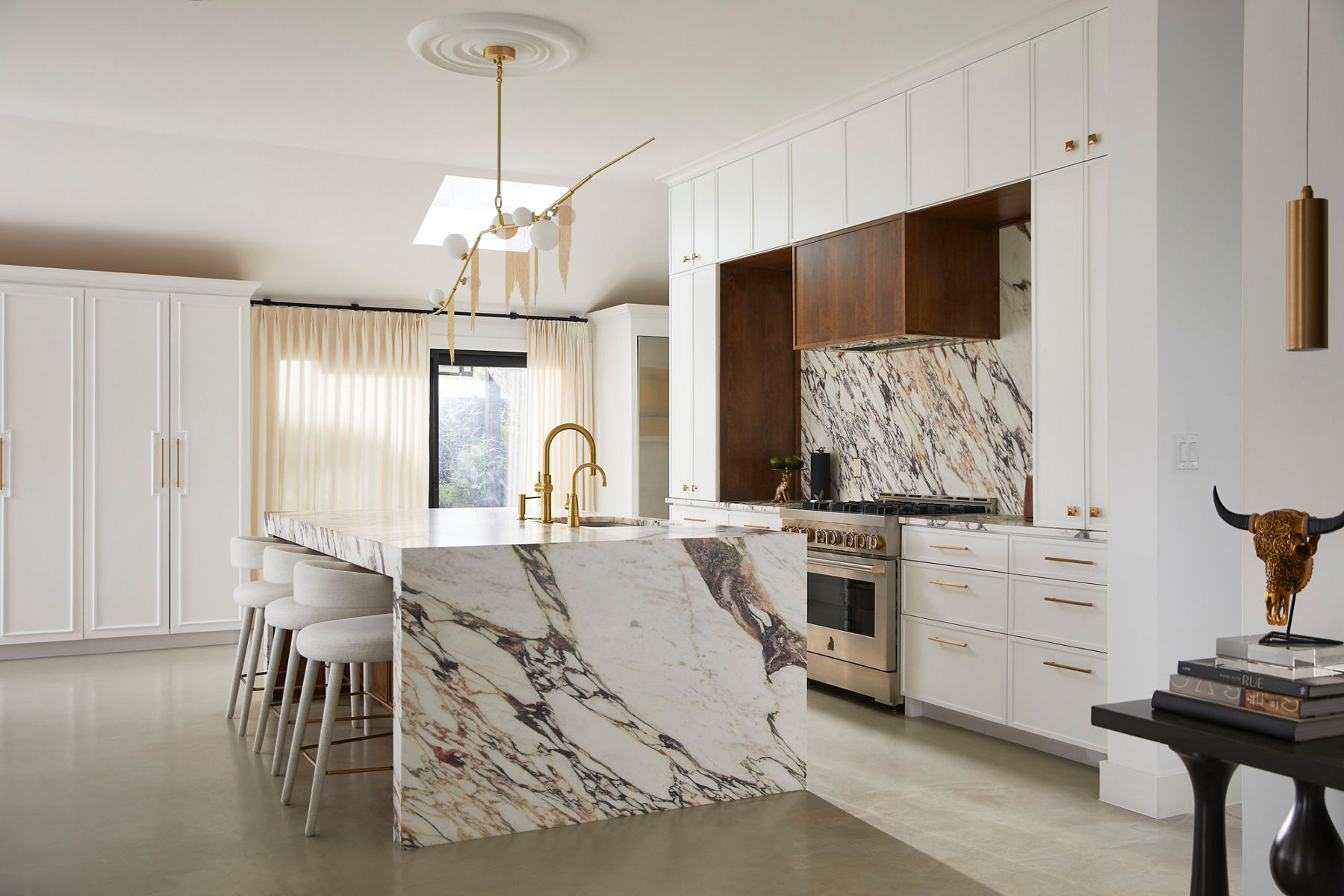 Wide image of Nikki's kitchen looking across the large kitchen island with marble counter top that wraps along the side to the floor. In the center of the island, under a chandelier, are two matching Kohler faucets in brass. One drinking water faucet and one full size Crue faucet.