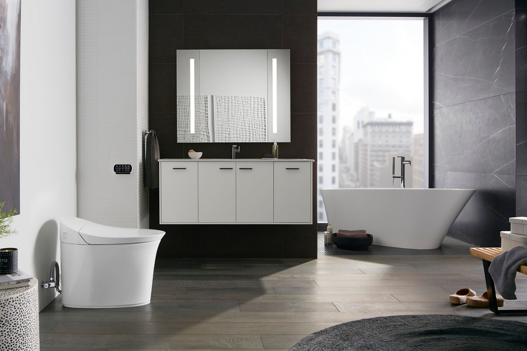A spacious bathroom with white KOHLER Veil Intelligent toilet, freestanding Veil bath, a white vanity with lighted mirror installed on a black wall.