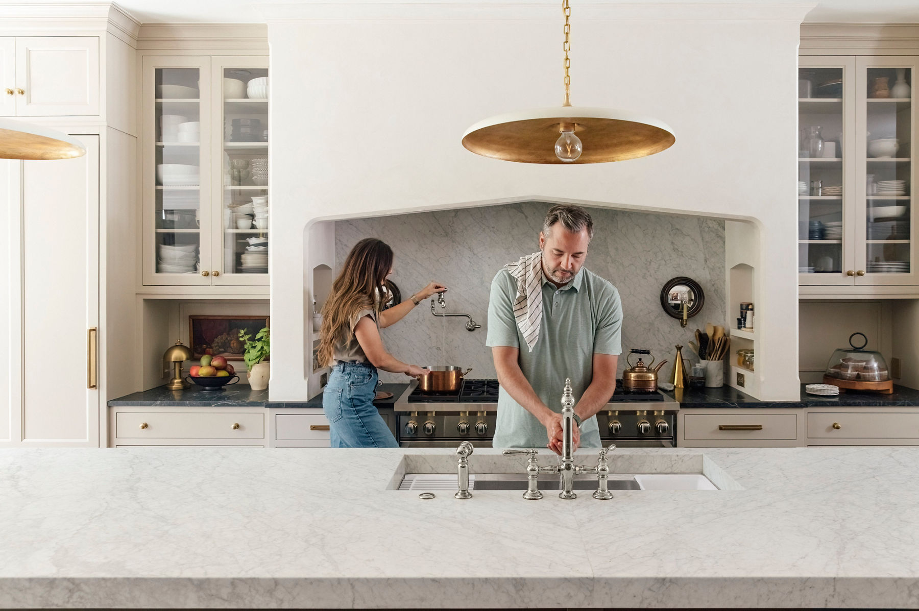 Wide image of Chris and Julia Marcum, the couple known online as ChrisLovesJulia, cooking in their large, open kitchen. Chris is standing behind the kitchen island sink with a towel over his shoulder and washing his hands in the workstation sink under the polished nickel Kohler Artifacts faucet, while Julia is standing at the stove behind him, filling a pot with water from the wall-mounted Kohler pot filler faucet.