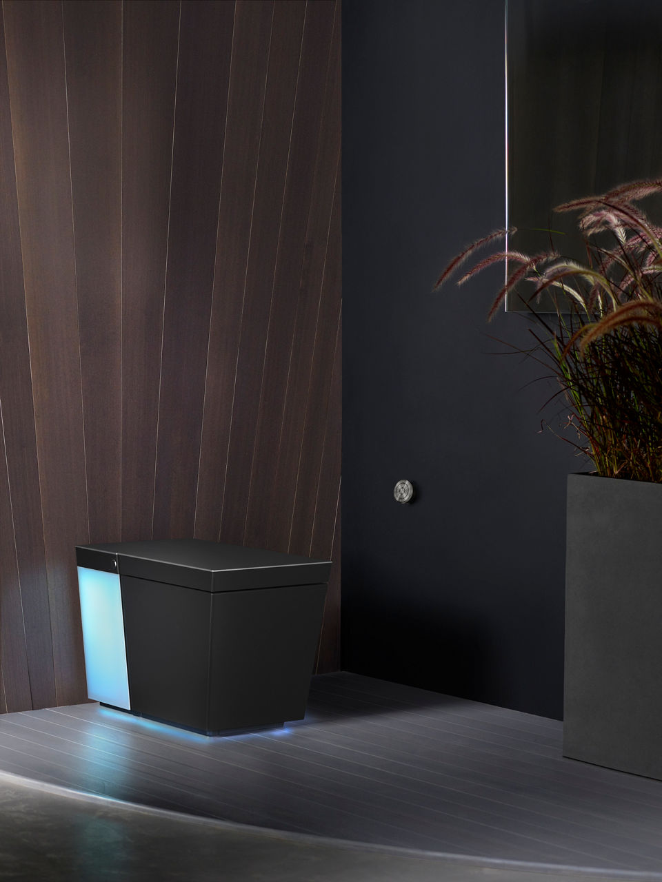 An illuminated Numi 2.0 toilet in a bathroom with gray floors, and brown wood and black painted walls.