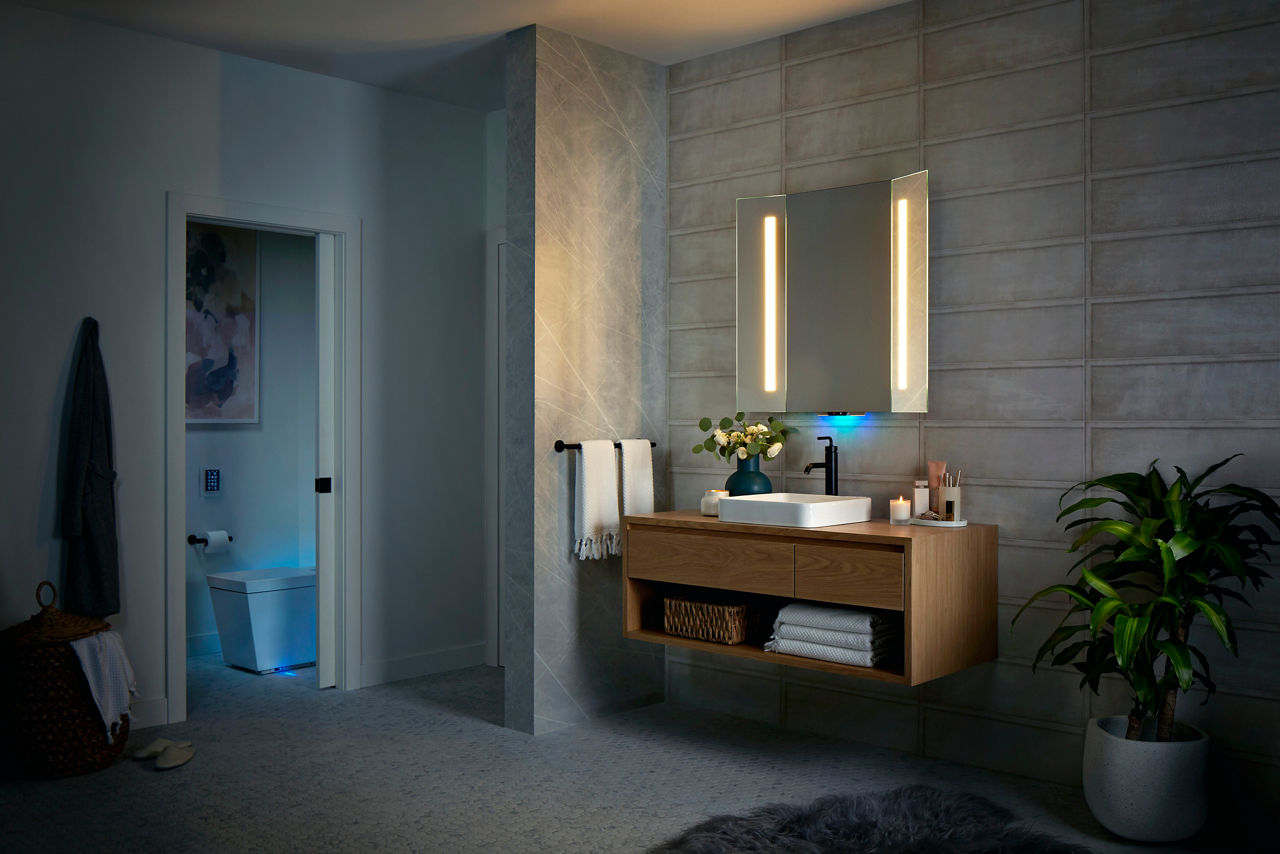The lights on a KOHLER NUMI intelligent toilet glow in a bathroom with a lighted mirror above a wall-mount vanity and white rectangle vessel sink.