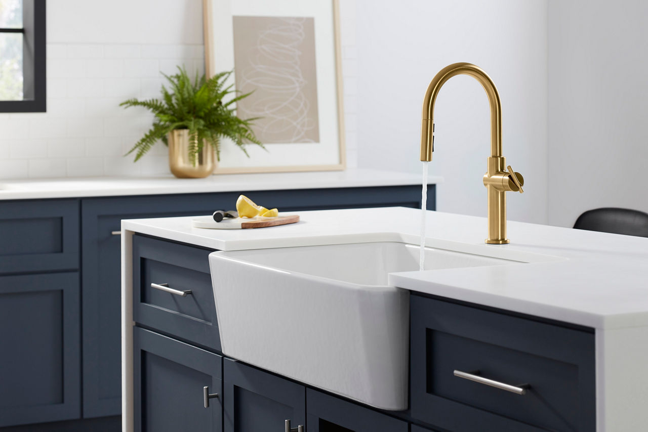 Water flows from a KOHLER Crue single lever handle faucet into a white farmhouse sink mounted in an island with blue cabinets and white countertops.