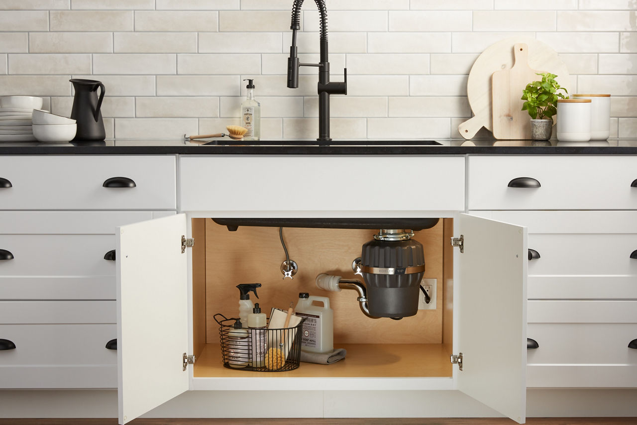 Doors to an under sink cabinet stand open, revealing a KOHLER garbage disposal and a basket of cleaning supplies.