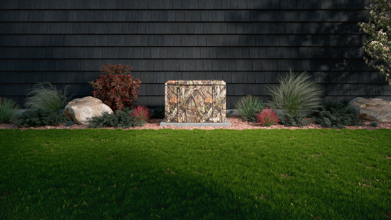 A camouflage covered home generator in front of a gray shingled house with grass in front.