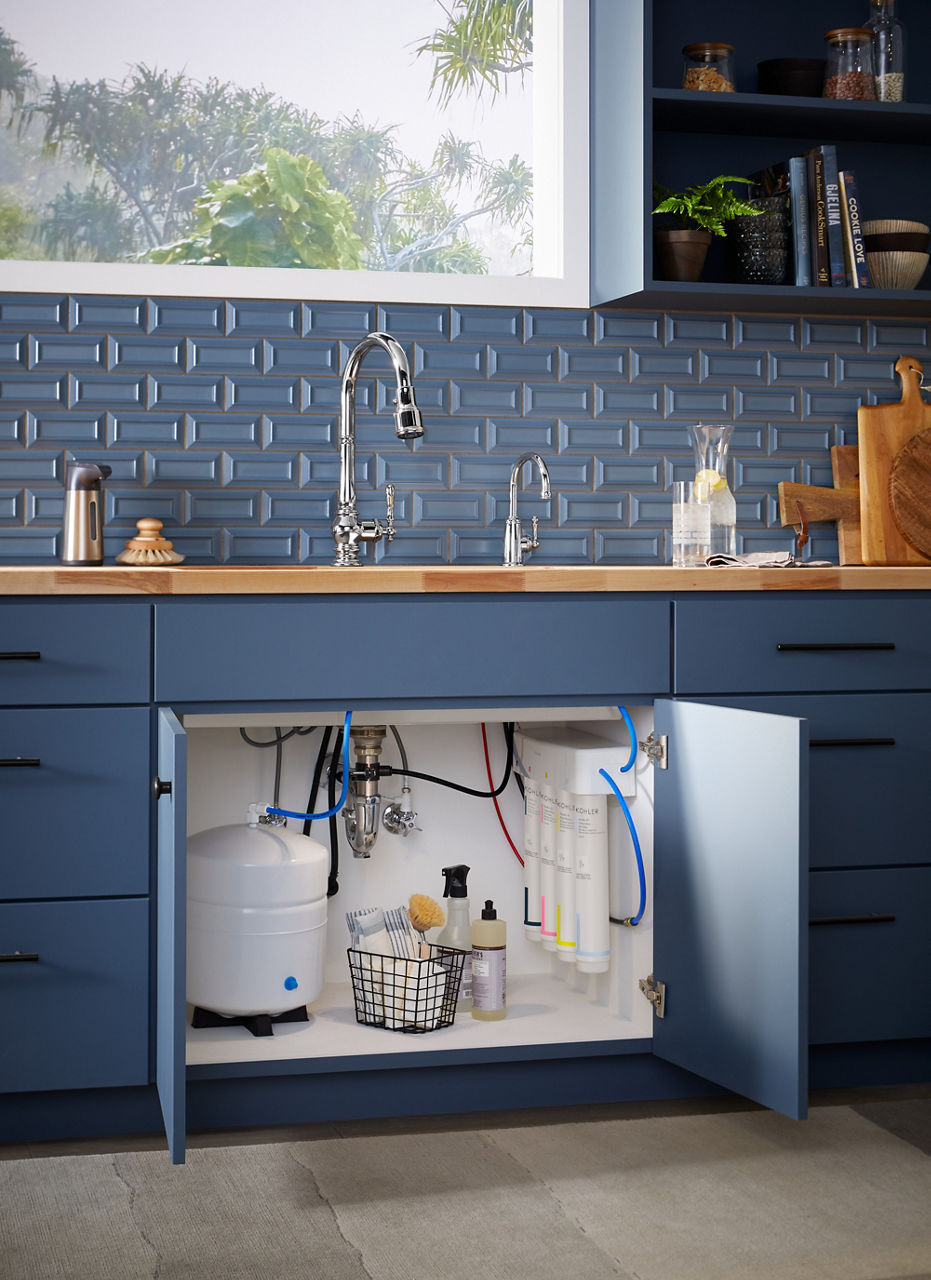 Doors to an under sink cabinet stand open, revealing a KOHLER Aquifer reverse osmosis water filtration system and a basket of cleaning products.