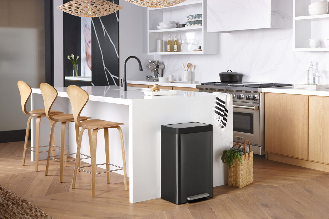 A Black Stainless KOHLER Step trashcan stands against a counter with white waterfall countertop.