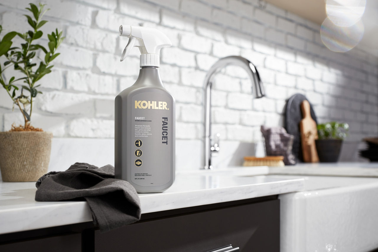 A spray bottle of KOHLER faucet cleaner and a towel sit on a white counter, next to a single handle kitchen faucet with tall arched spout.