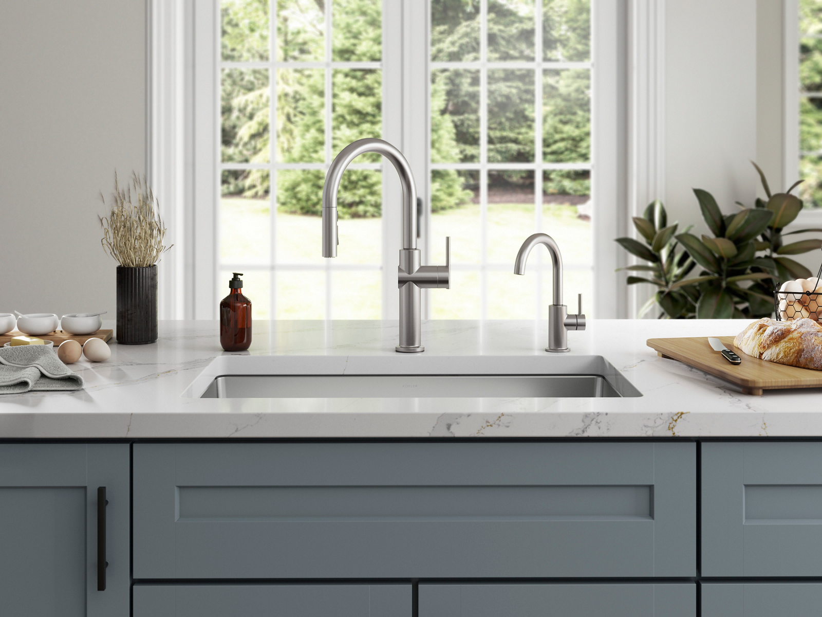A close up shot of a sunny kitchen with a kitchen sink at center. The sink is attached to a white counter top and designed with a silver faucet with a silver water tap faucet.