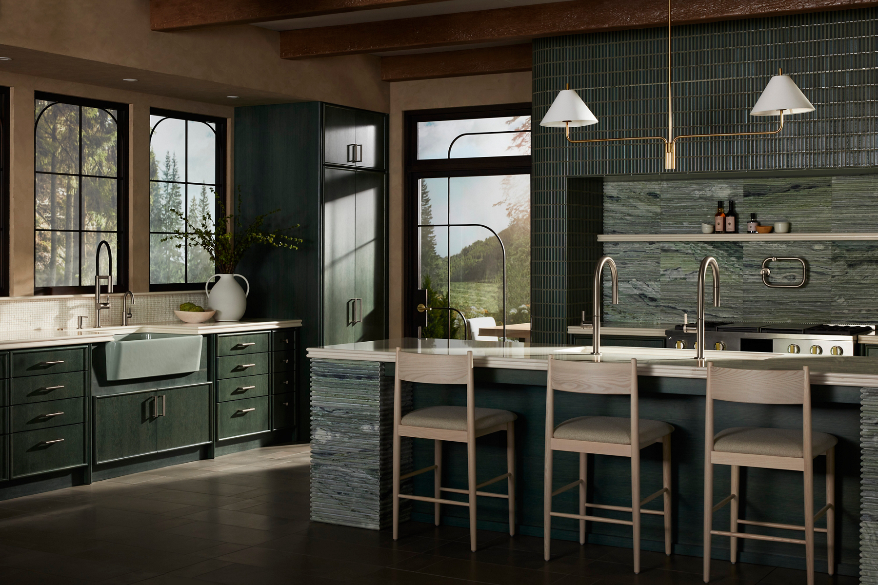 A wide lifestyle image of a sundrenched kitchen designed with dark moss green tiling and beige textured walls. There's a kitchen island at focus with a double silver sink faucet faced away from the camera. Behind the island is a kitchen counter with a silver stove and decorative shelf above. To the leftside of the image is another kitchen sink designed with a muted mint green body and silver faucets.