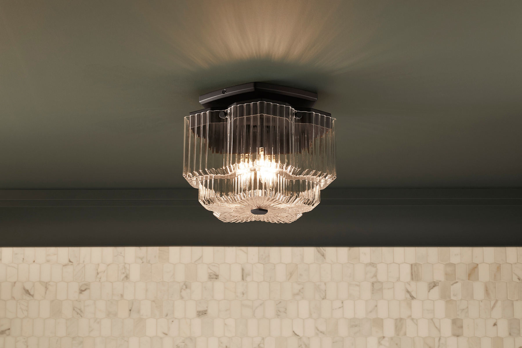 A KOHLER Occasions flush-mount light fixture with ridged glass shade casts a rippled reflection on a dark gray ceiling.