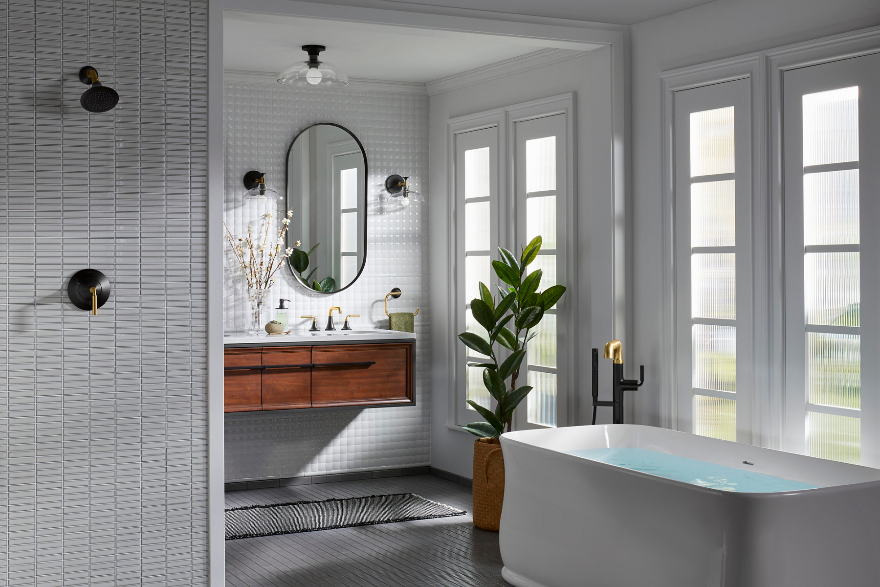 A KOHLER white freestanding bath full of water and ready for a soak in a light and bright bathroom with white tiled shower, wall hung vanity, and capsule mirrors.