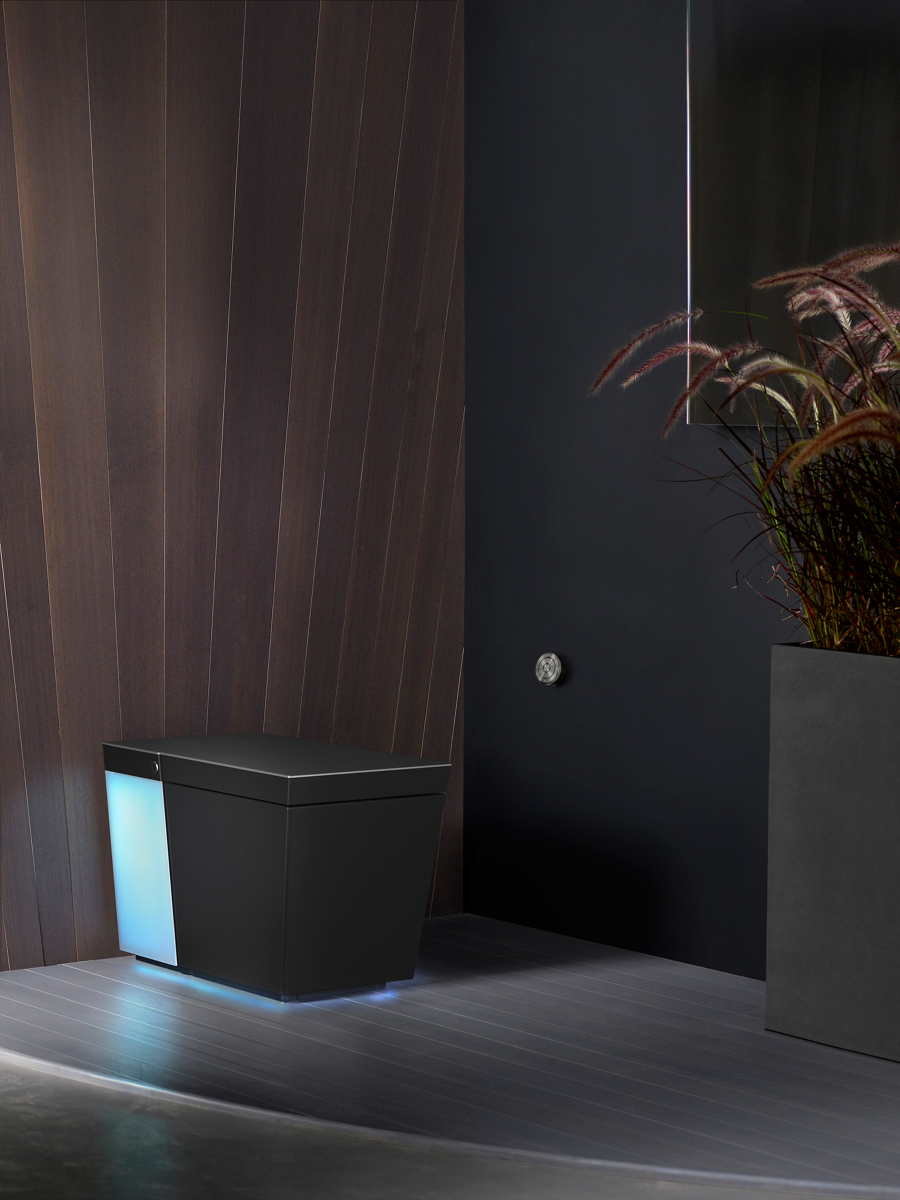 A black Numi 2.0 toilet with the underside and back illuminated in a bathroom with gray floors, a slatted wood wall behind it and a black wall with the toilet remote control to its side.