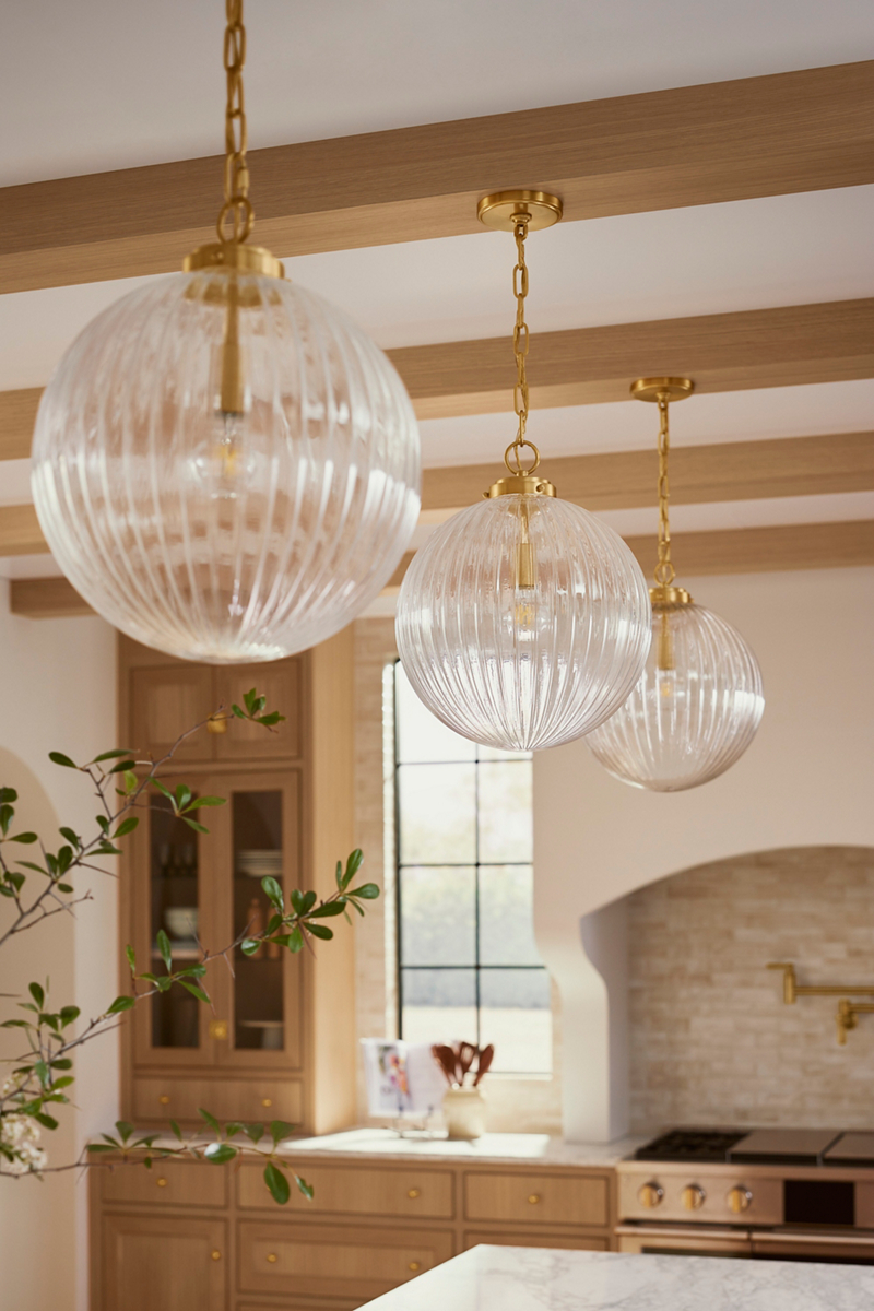 Three brushed brass and reeded globe glass pendant lights designed by Shea McGee hanging from a wood-beamed ceiling.