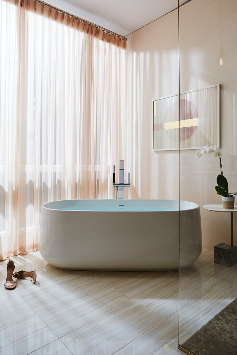 Light streams through sheer curtains and illuminates a white oval freestanding bath with floor mounted bath filler.