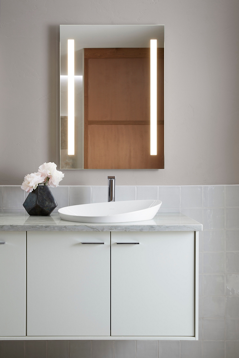 A KOHLER Verdera lighted mirror above a white Veil vessel sink and gray vanity top with a black vase of white peonies.